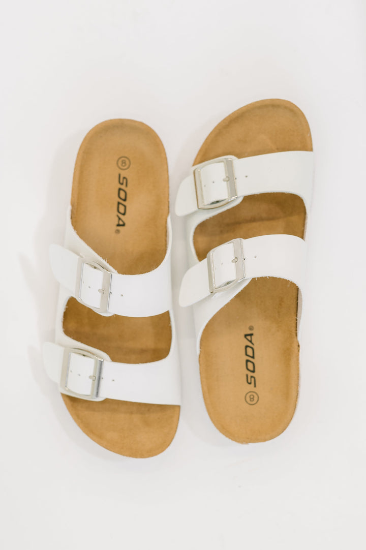 Top-down flat lay of white double-buckle slide sandals by Soda with a tan cork footbed, on a white background.