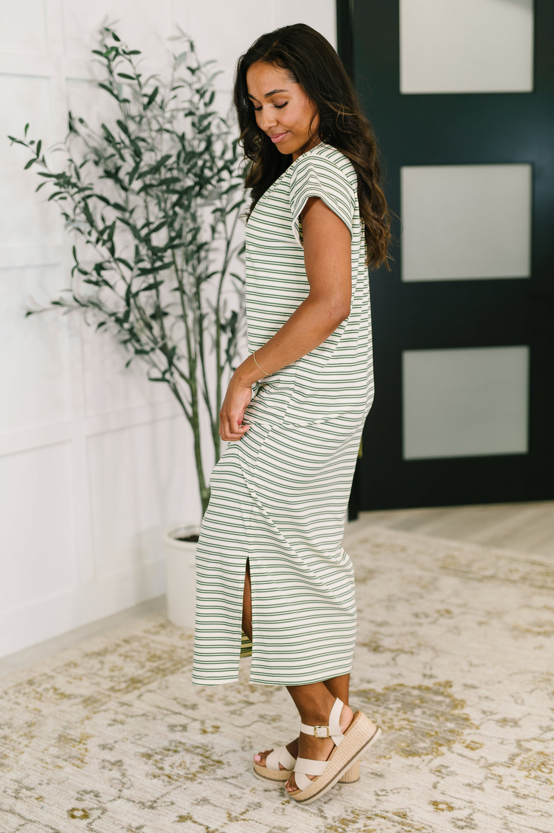 Side/back view of a woman in a white and olive green striped short-sleeve maxi dress with a side slit, looking down, paired with cream platform wedge sandals, posed in front of a potted olive tree and dark French door