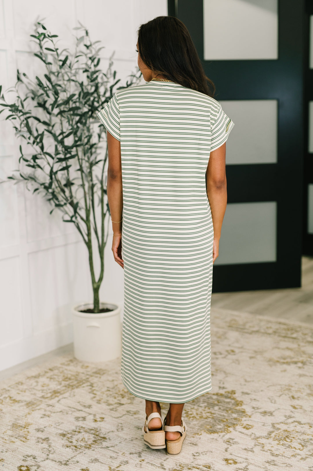 Back view of a woman in a white and olive green striped short-sleeve maxi dress, paired with cream platform wedge sandals, posed in front of a potted olive tree and dark French door