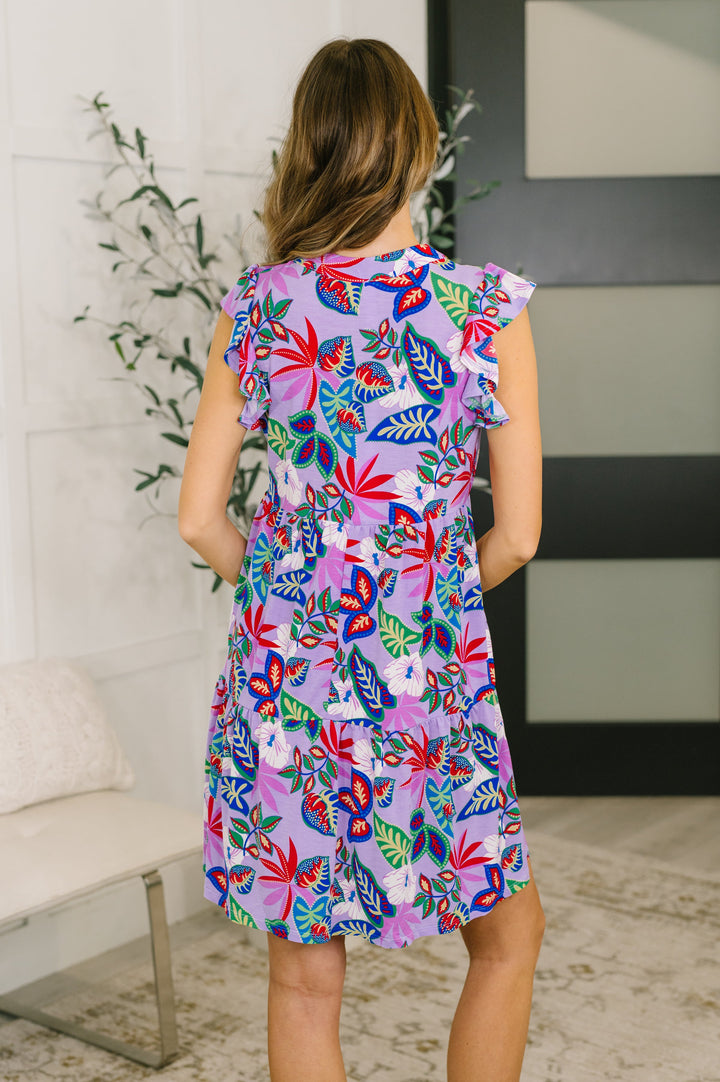 Back view of a woman wearing a lavender tropical floral mini dress with flutter sleeves, posed in front of a white bench and leafy plant