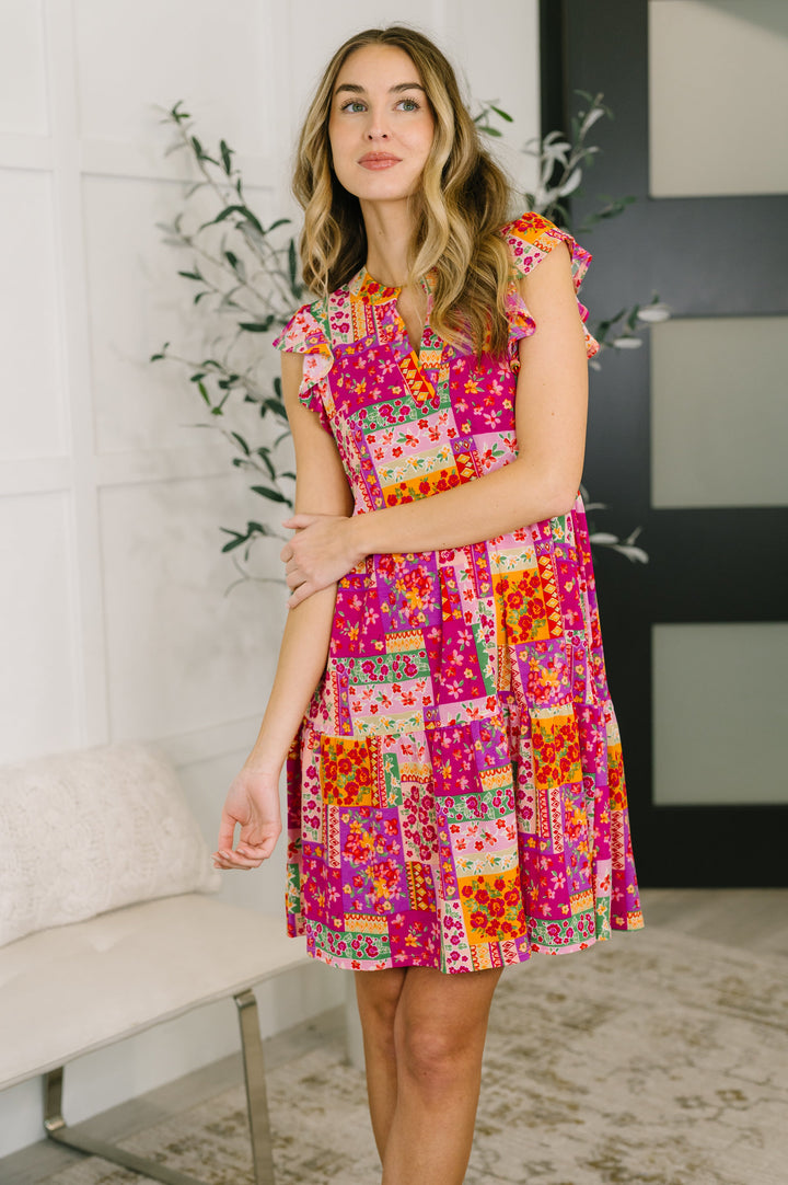 Woman wearing a bright pink patchwork floral mini dress with flutter sleeves, posed in front of a white bench and leafy plant, full-length view