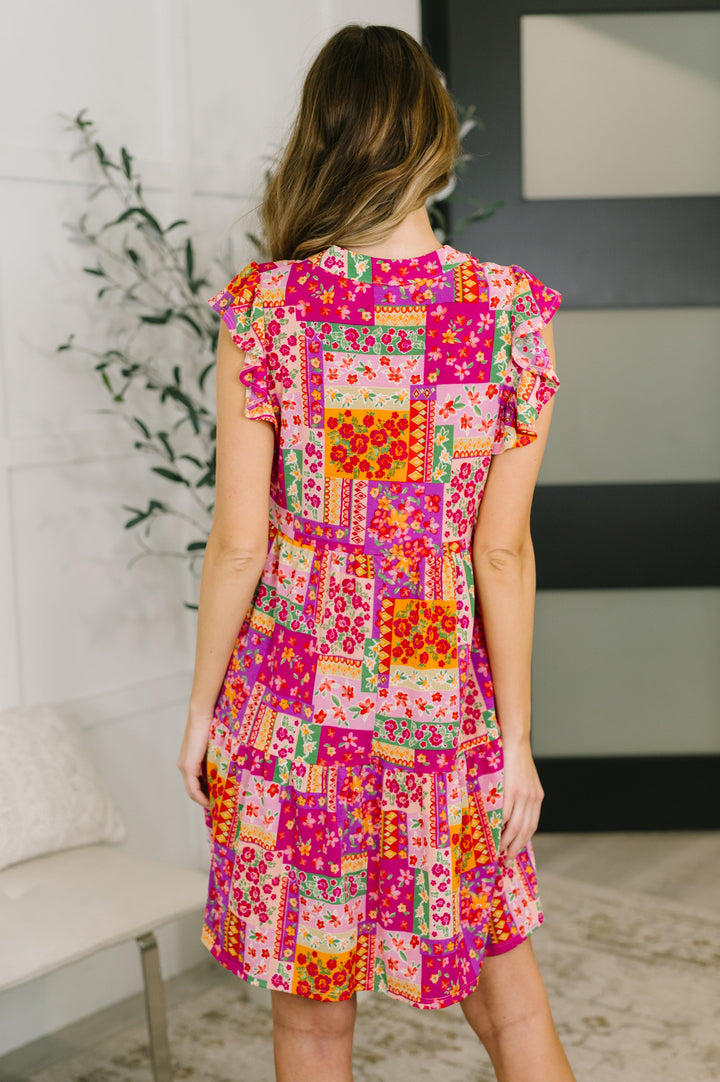 Back view of a woman wearing a bright pink patchwork floral mini dress with flutter sleeves, posed in front of a white bench and leafy plant