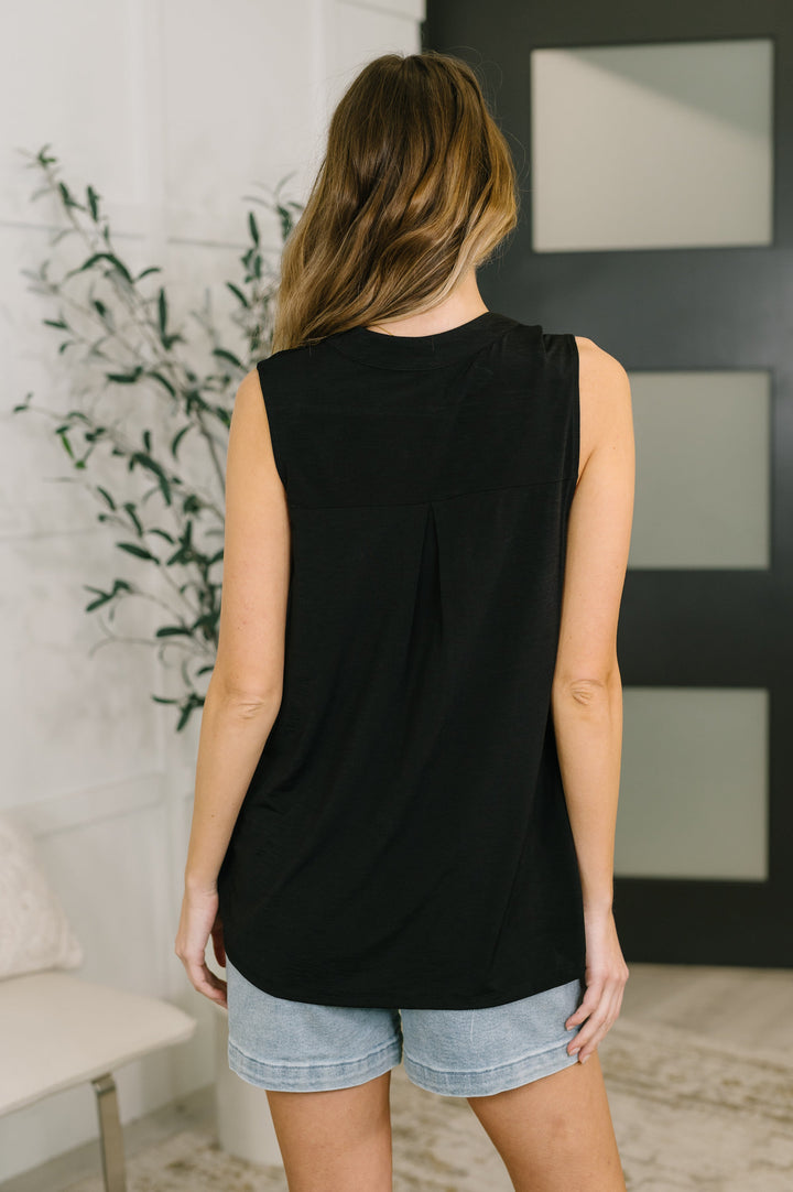 Back view of a woman in a black sleeveless split-neck top with a pleat detail, paired with wide-leg jeans.