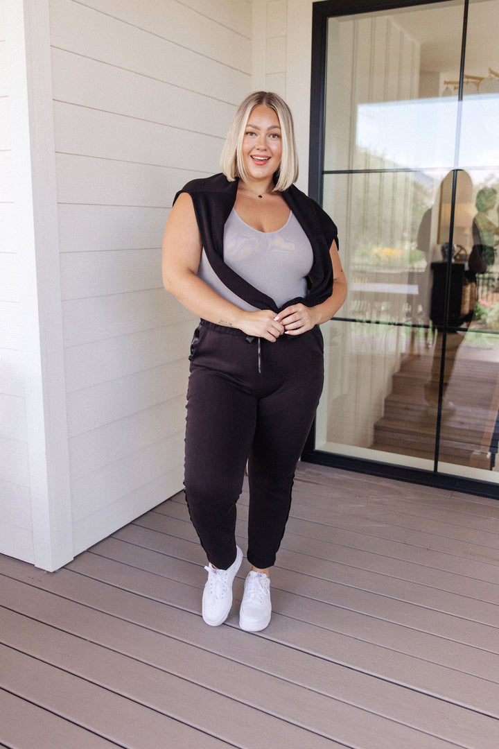Plus size model smiling in a gray scoop-neck bodysuit with a black sleeveless vest, black tapered jogger pants, and white sneakers, standing in front of a white shiplap wall and black French door