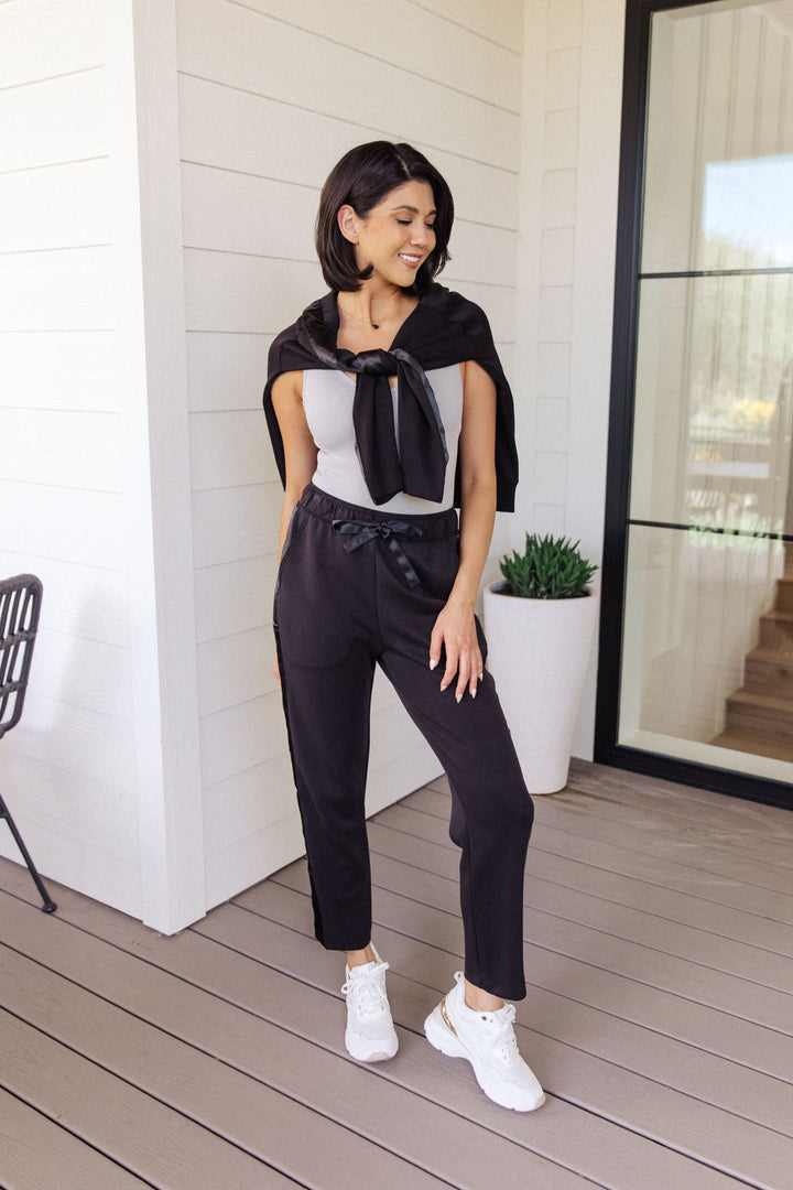 Woman smiling in a gray scoop-neck bodysuit with wide-leg rhinestone jeans and white sneakers, standing in front of black French doors with a potted plant