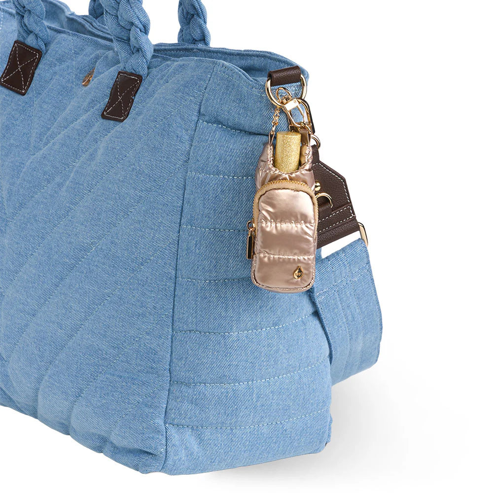 Champagne gold metallic quilted puffer lip balm keychain holder clipped onto the side of a light blue denim quilted tote bag with braided handles.