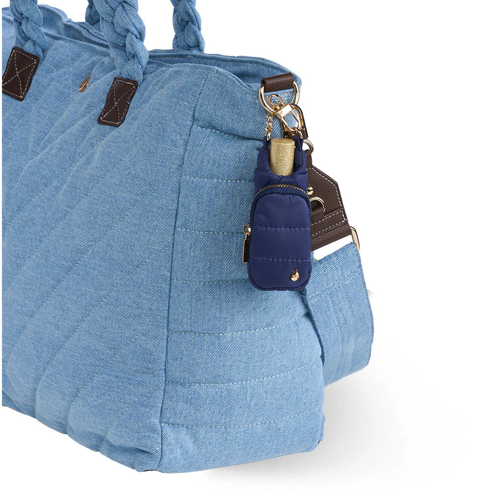 Navy blue quilted puffer lip balm keychain holder clipped onto the side of a light blue denim quilted tote bag with braided handles.