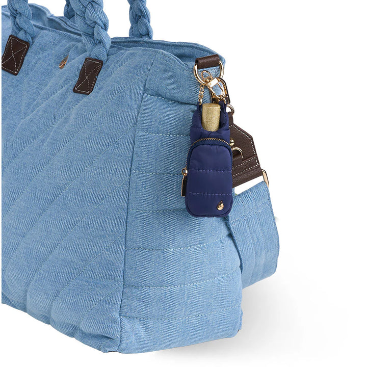 Navy blue quilted puffer lip balm keychain holder clipped onto the side of a light blue denim quilted tote bag with braided handles.