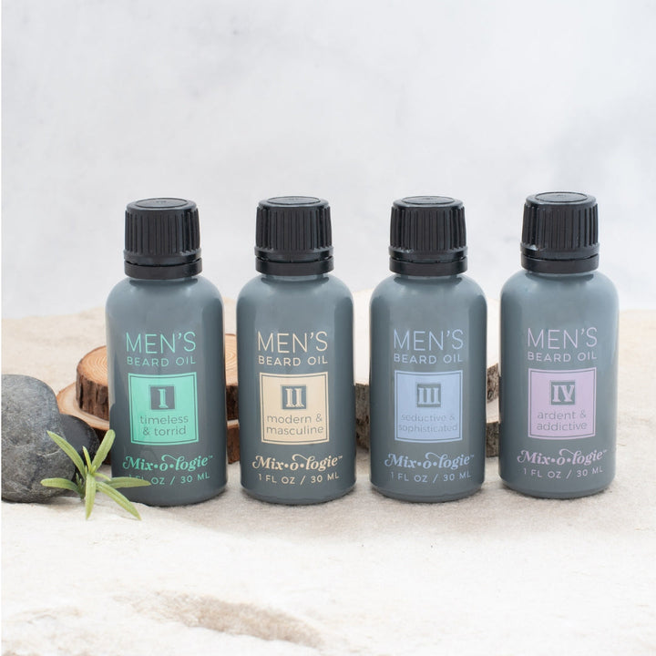 Mixologie Men's Beard Oil collection, four 1 fl oz slate gray bottles — I Timeless & Torrid, II Modern & Masculine, III Seductive & Sophisticated, and IV Ardent & Addictive — displayed on sandy surface with wood slices, river rocks, and green sprig