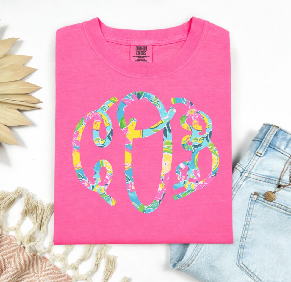 Folded hot pink Comfort Colors t-shirt with a large circular floral monogram graphic in turquoise, yellow, green, and pink