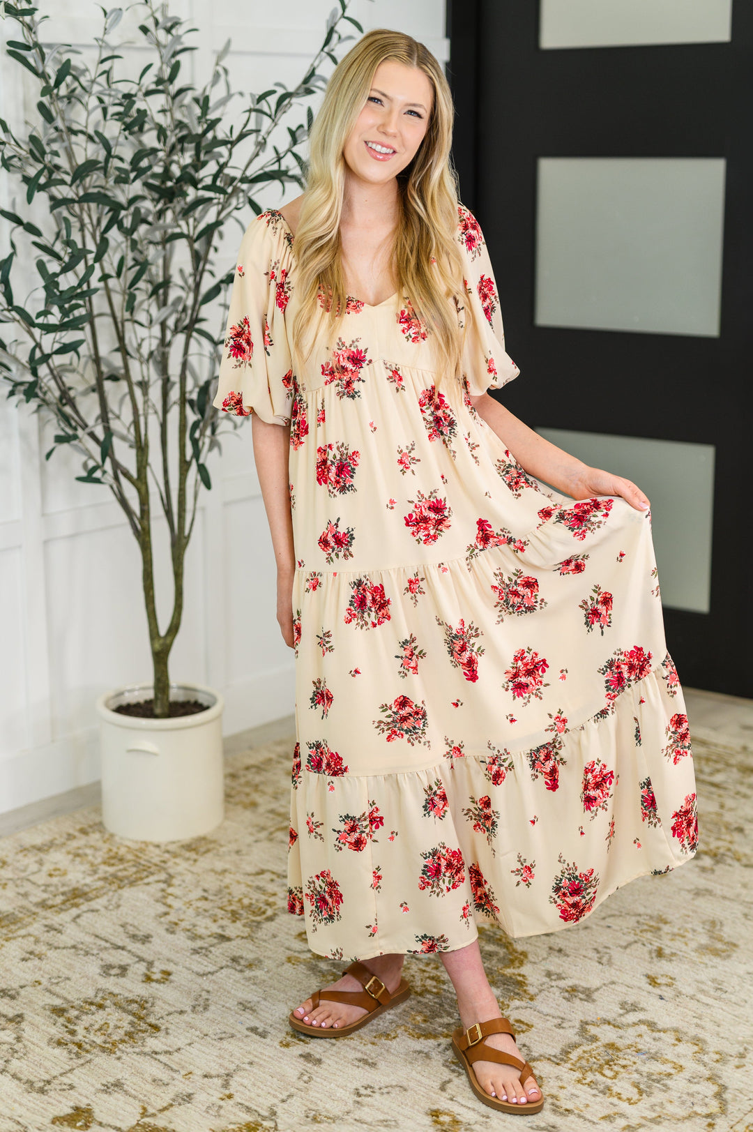 Full-length front view of a woman smiling in a cream floral midi dress with red roses, puff sleeves, and a tiered skirt, paired with tan buckle sandals