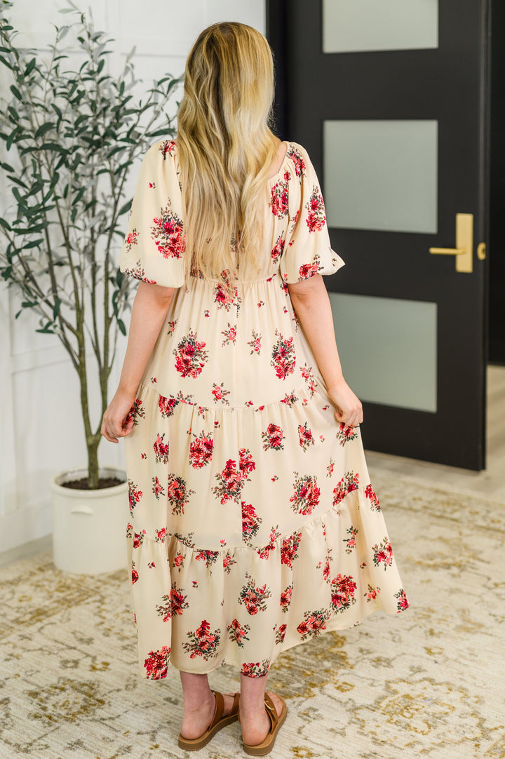 Back view of a woman in the cream red floral puff-sleeve tiered midi dress with tan buckle sandals.