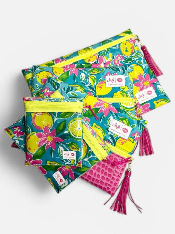 Makeup Junkie Bags teal lemon and floral print collection including large clutch, medium clutch, small pouch, and pink croc mini clutch with yellow zippers and pink tassels on light gray background