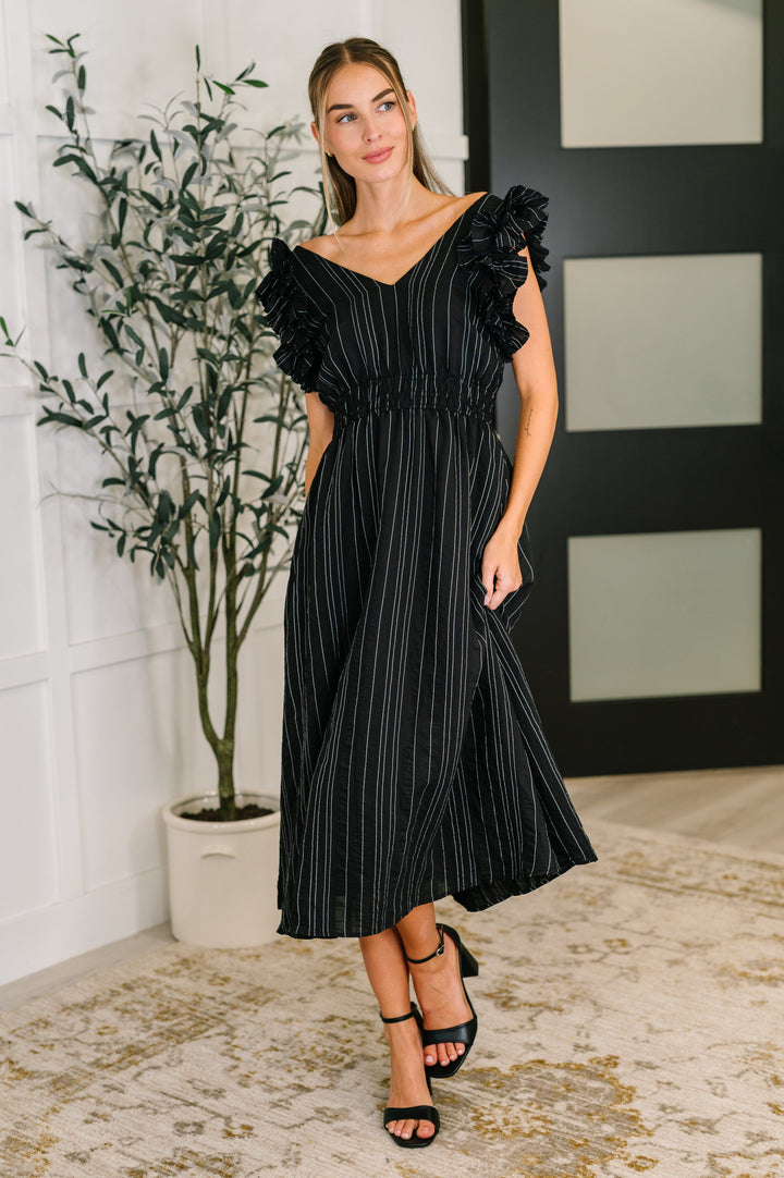 Woman wearing a black metallic stripe midi dress with ruffle flutter sleeves and smocked waist, styled with black block heel sandals.