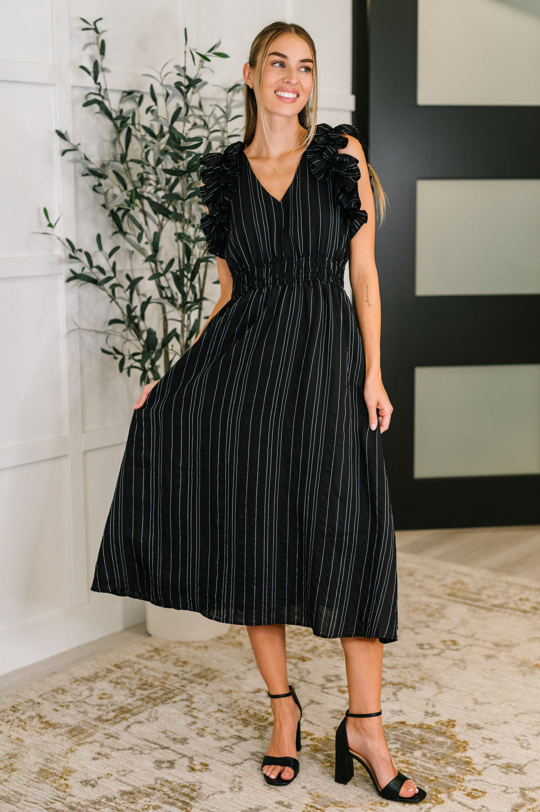 Woman wearing a black metallic stripe midi dress with ruffle flutter sleeves, V-neckline, and smocked waist, styled with black block heel sandals.