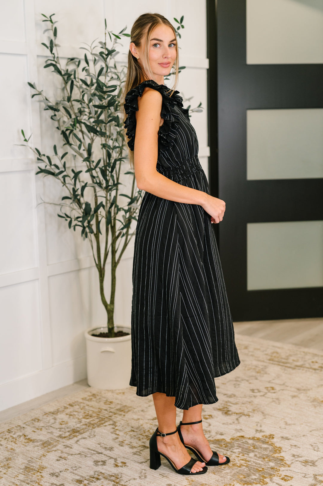 Side view of woman wearing a black metallic stripe midi dress with ruffle flutter sleeves and A-line midi skirt, styled with black block heel sandals.