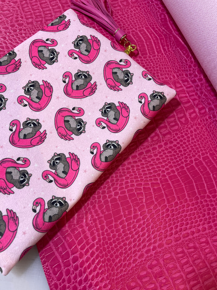 Makeup Junkie Bags cosmetic bag with raccoon on flamingo float print and pink tassel zipper pull, shown alongside hot pink crocodile-embossed and metallic pink surfaces