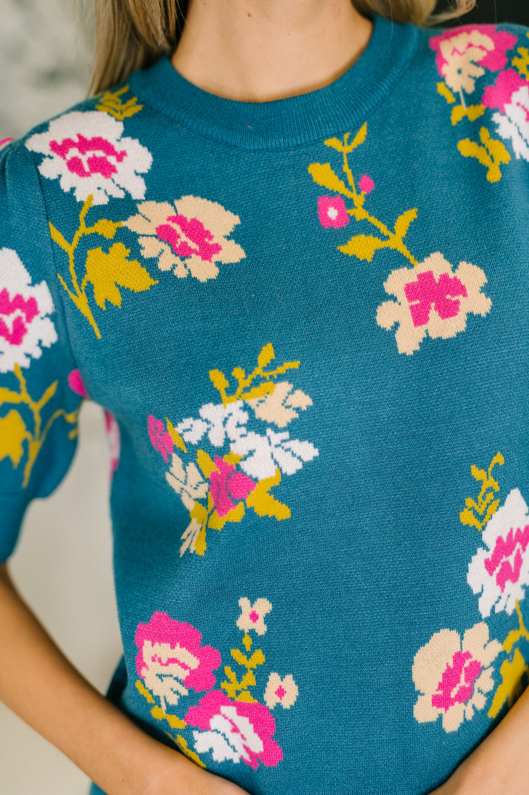 Close-up of teal floral jacquard sweater top showing pink, white, and yellow flower pattern and textured knit detail.
