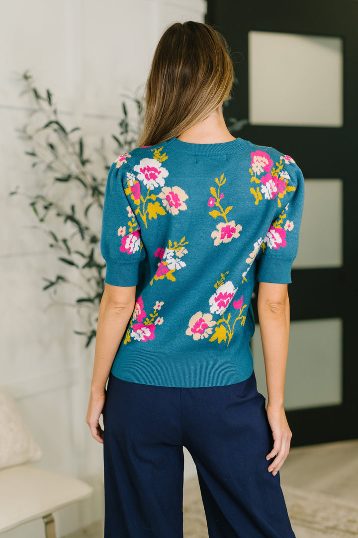 Back view of teal short-sleeve floral jacquard sweater top showing allover pink, white, and yellow floral pattern and smocked hem, styled with navy wide-leg pants.