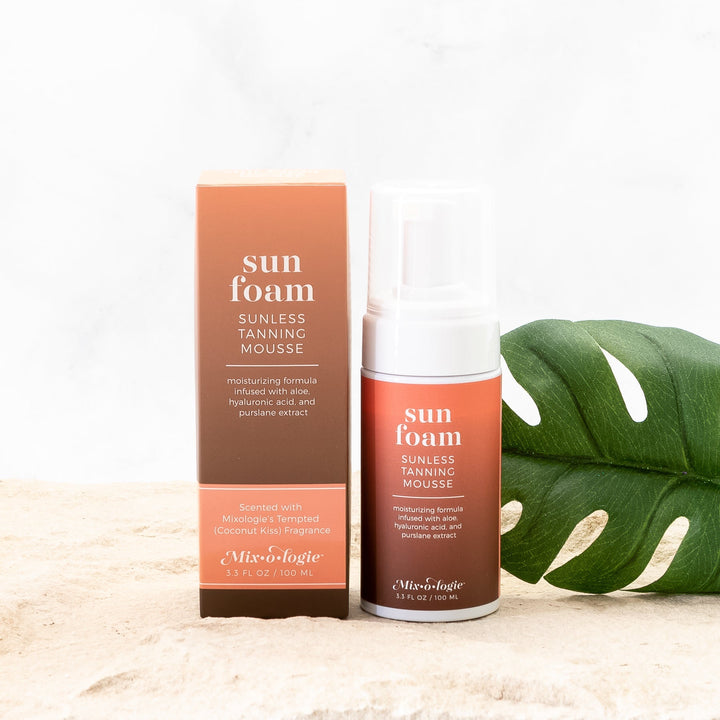 Mixologie Sun Foam Sunless Tanning Mousse 3.3 fl oz full-size bottle with matching brown and orange box styled on sandy surface with monstera leaf