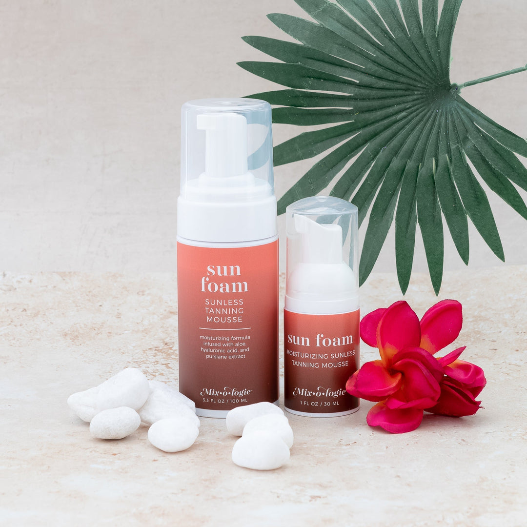Mixologie Sun Foam Sunless Tanning Mousse full-size 3.3 fl oz and travel 1 fl oz bottles styled on sandy surface with white river rocks, red plumeria flower, and palm leaf