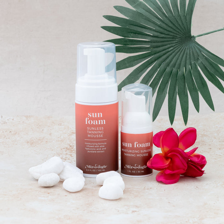 Mixologie Sun Foam Sunless Tanning Mousse full-size 3.3 fl oz and travel 1 fl oz bottles styled on sandy surface with white river rocks, red plumeria flower, and palm leaf