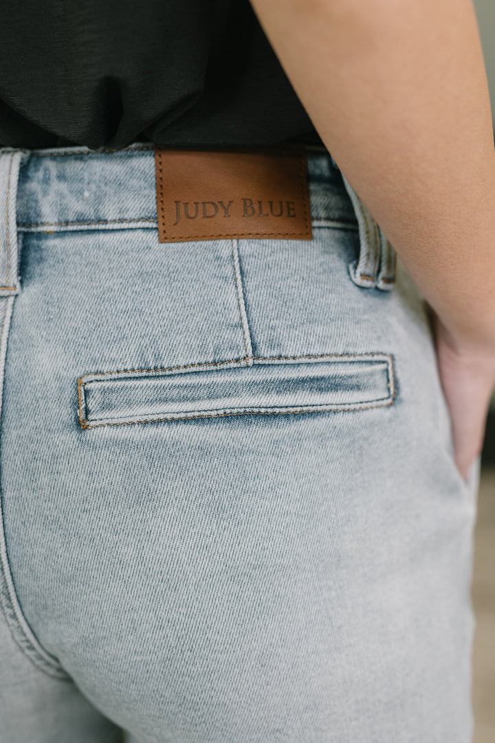 Close-up of light wash Judy Blue denim midi shorts showing leather brand patch, back welt pocket, and belt loop detail.