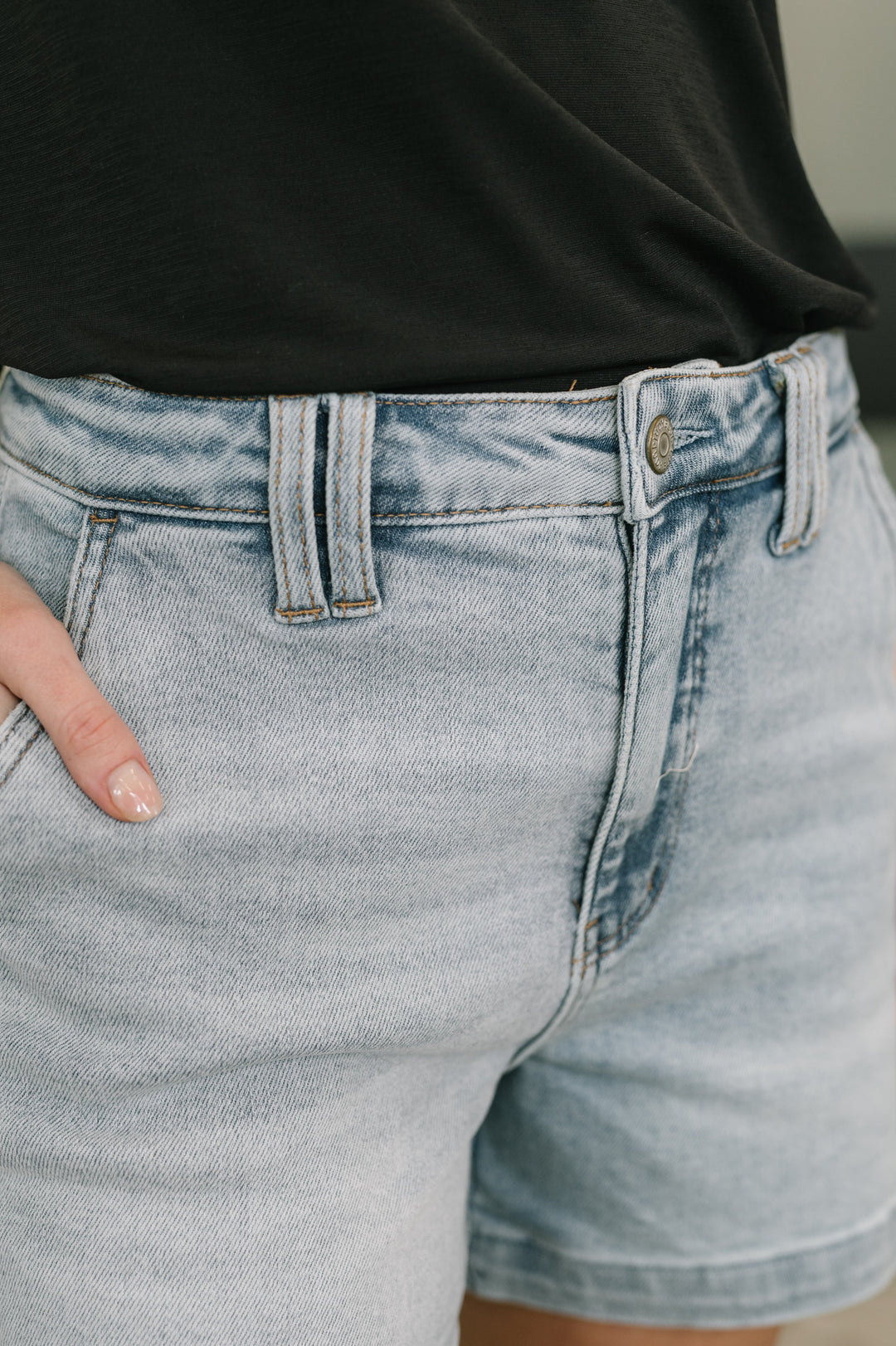 Close-up of light wash high-rise denim midi shorts showing button closure, belt loops, and front pocket detail.
