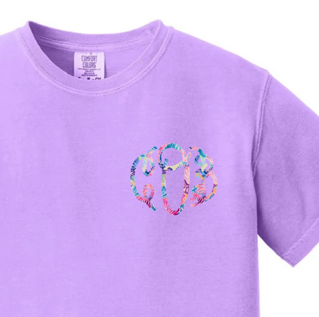 Lavender Comfort Colors t-shirt with a circular script monogram graphic in a colorful tropical floral pattern on the left chest