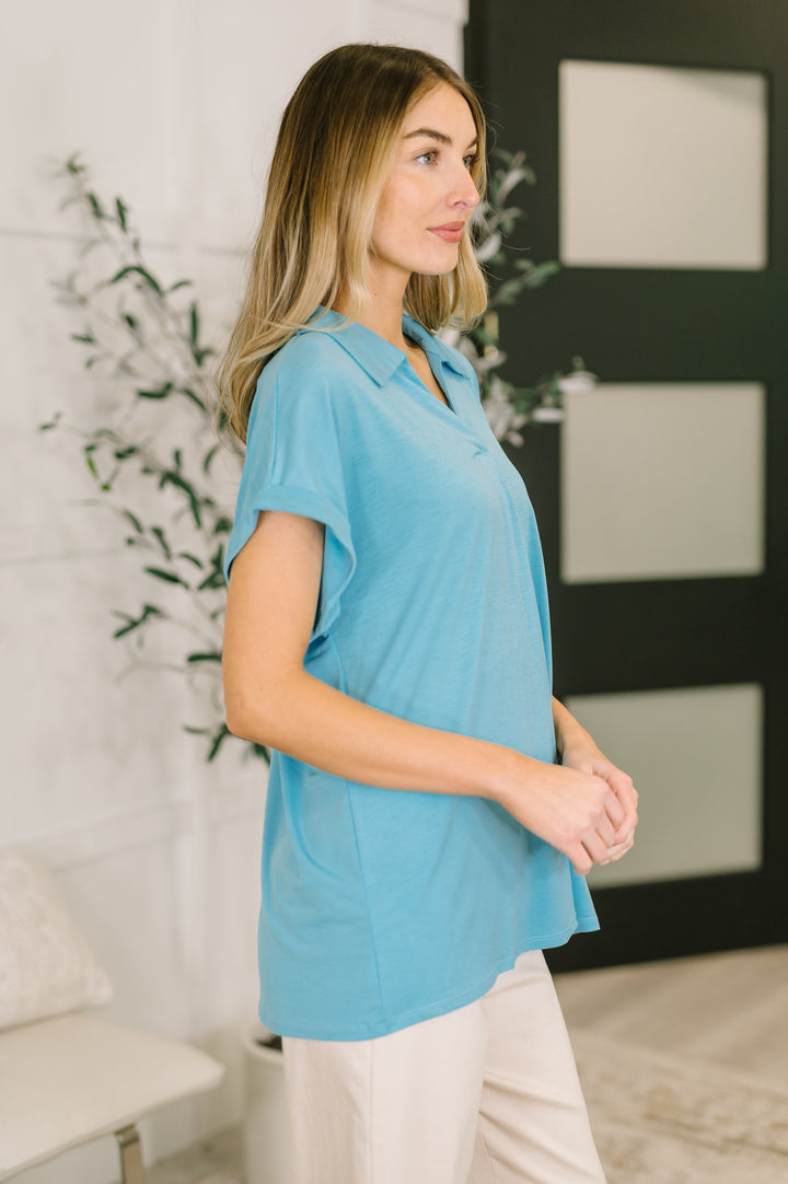 Side view of woman wearing a sky blue short-sleeve collared polo top showing relaxed silhouette and curved hem, styled with cream wide-leg pants.