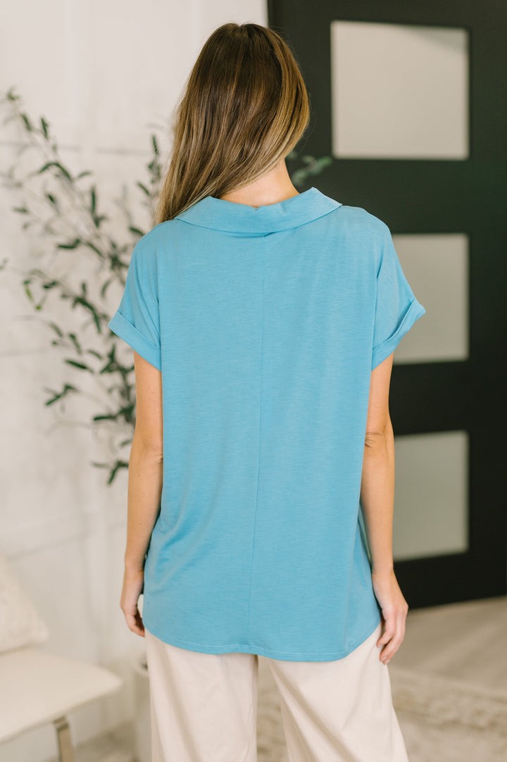 Back view of sky blue short-sleeve collared polo top showing clean seaming, cuffed sleeves, and curved hem, styled with cream wide-leg pants.