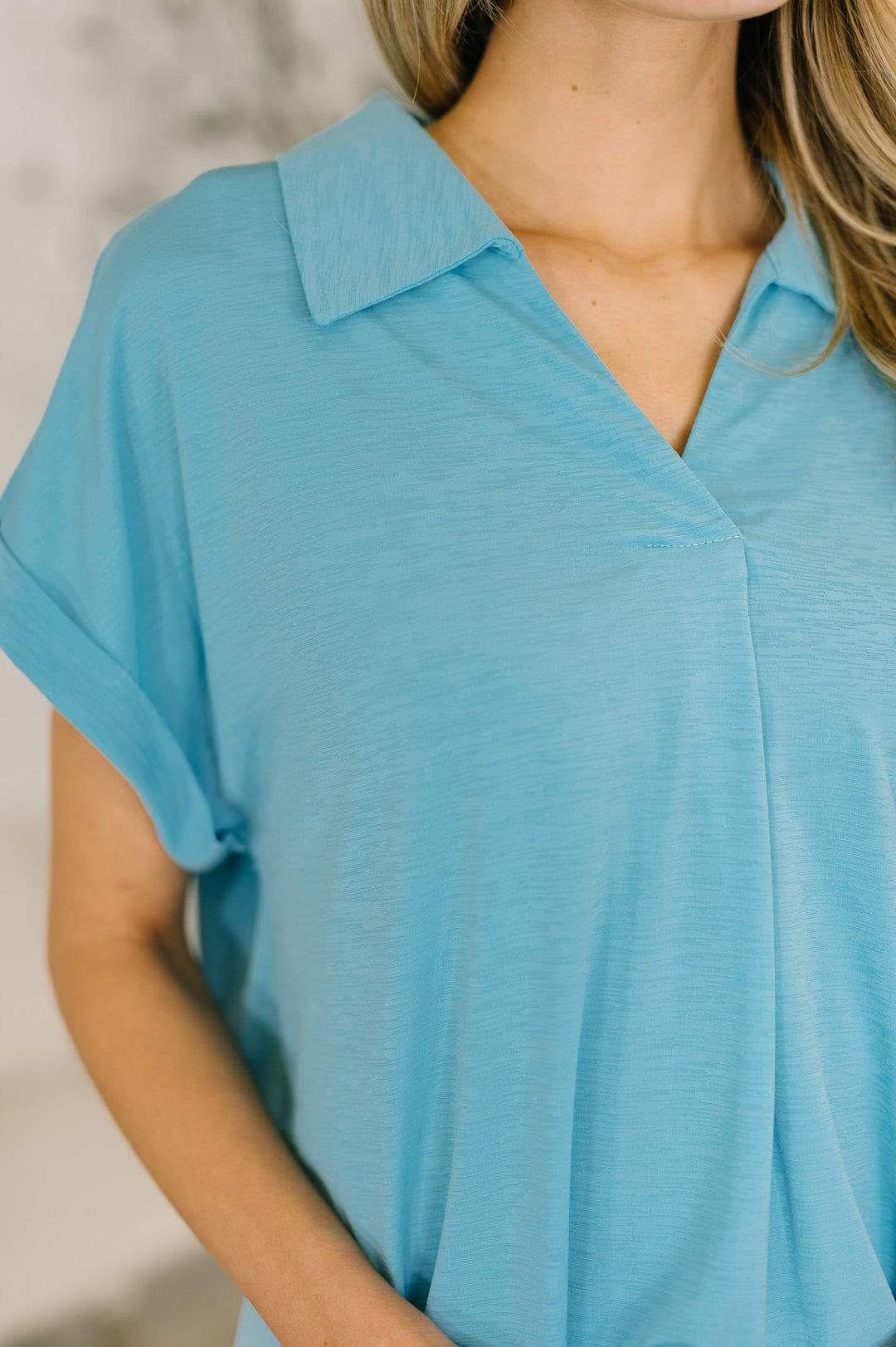 Close-up of sky blue short-sleeve collared polo top showing V-neckline, collar detail, center pleat, and cuffed sleeve texture.