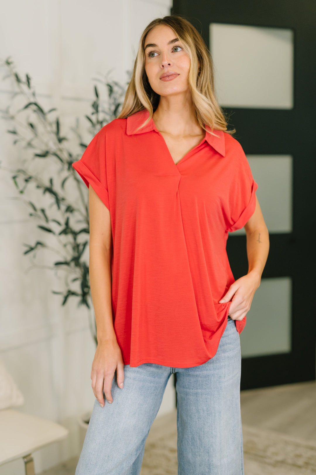 Woman wearing a coral red short-sleeve collared polo top with V-neckline and cuffed sleeves, styled with wide-leg light wash jeans.
