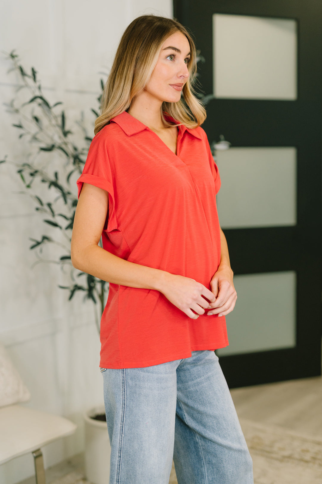 Side view of woman wearing a coral red short-sleeve collared polo top showing relaxed silhouette and curved hem, styled with wide-leg light wash jeans.