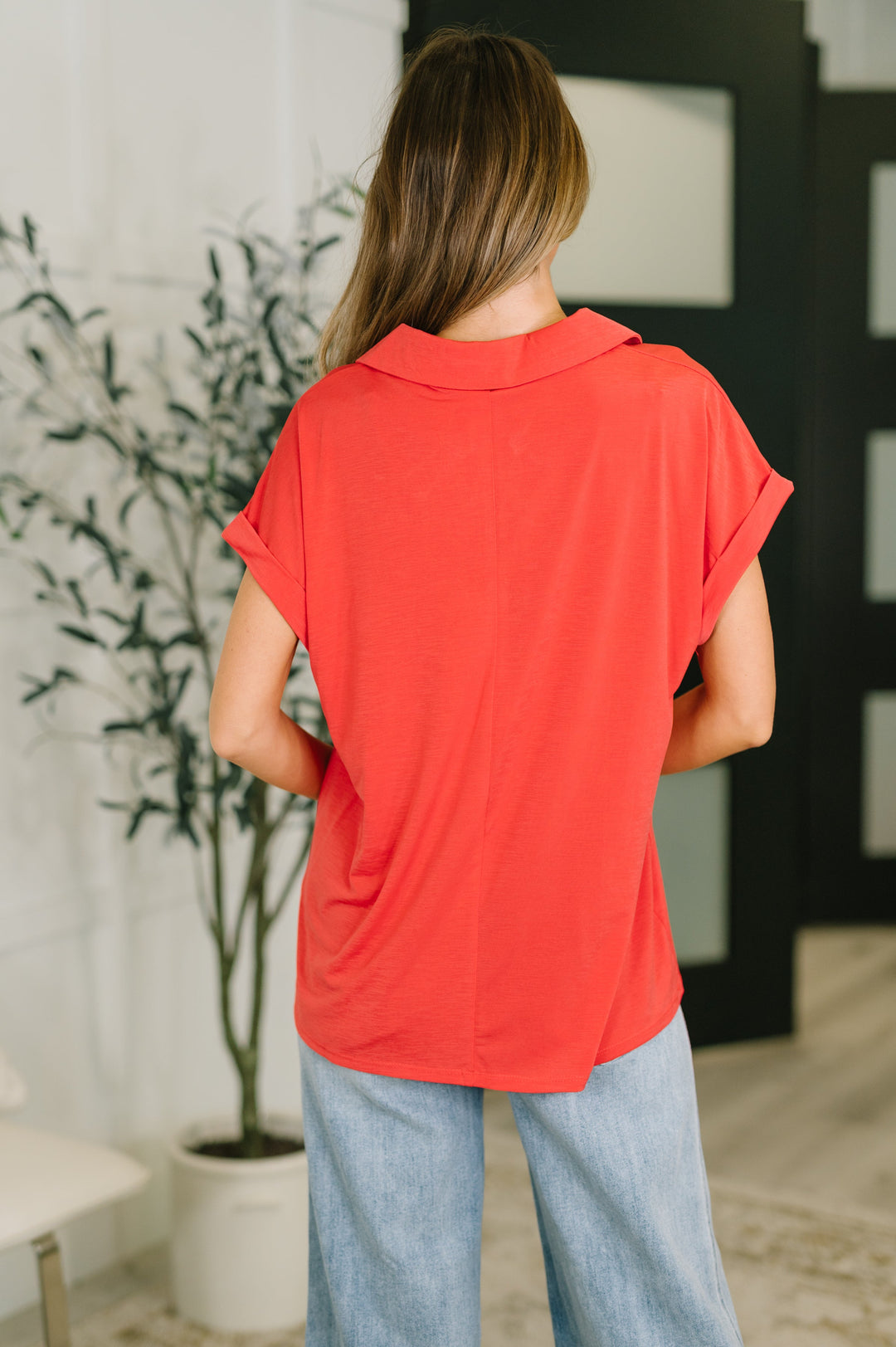Back view of coral red short-sleeve collared polo top showing clean seaming, cuffed sleeves, and curved hem, styled with wide-leg light wash jeans.