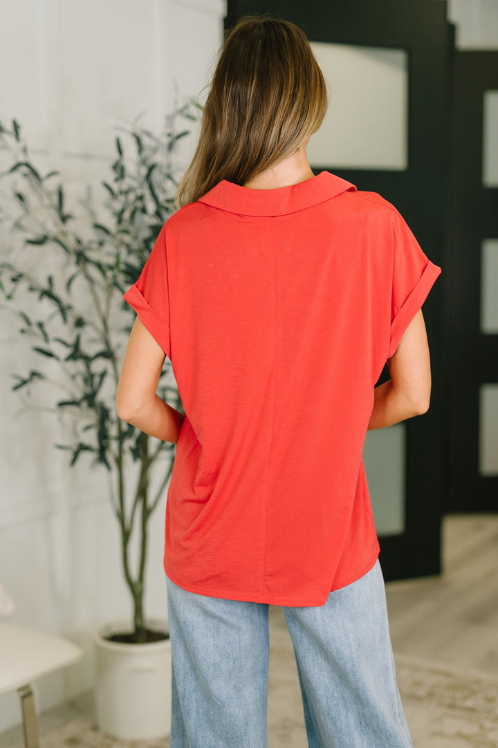 Back view of coral red short-sleeve collared polo top showing clean seaming, cuffed sleeves, and curved hem, styled with wide-leg light wash jeans.