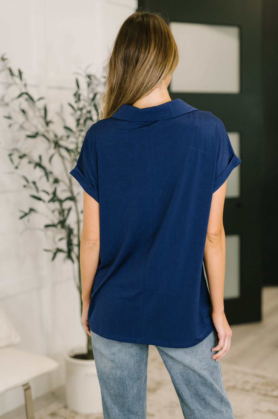 Back view of navy short-sleeve collared polo top showing clean seaming and curved hem, styled with light wash jeans.

