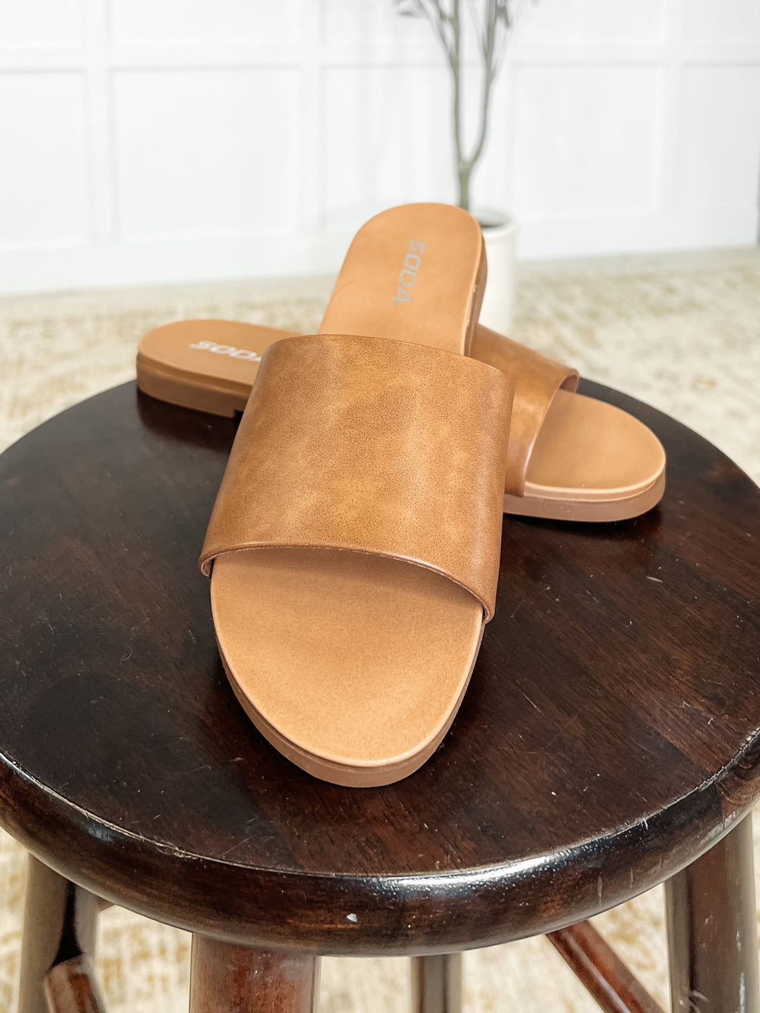 Close-up top view of tan leather single-band slide sandals on a dark wood stool, showing the wide strap and cushioned footbed detail
