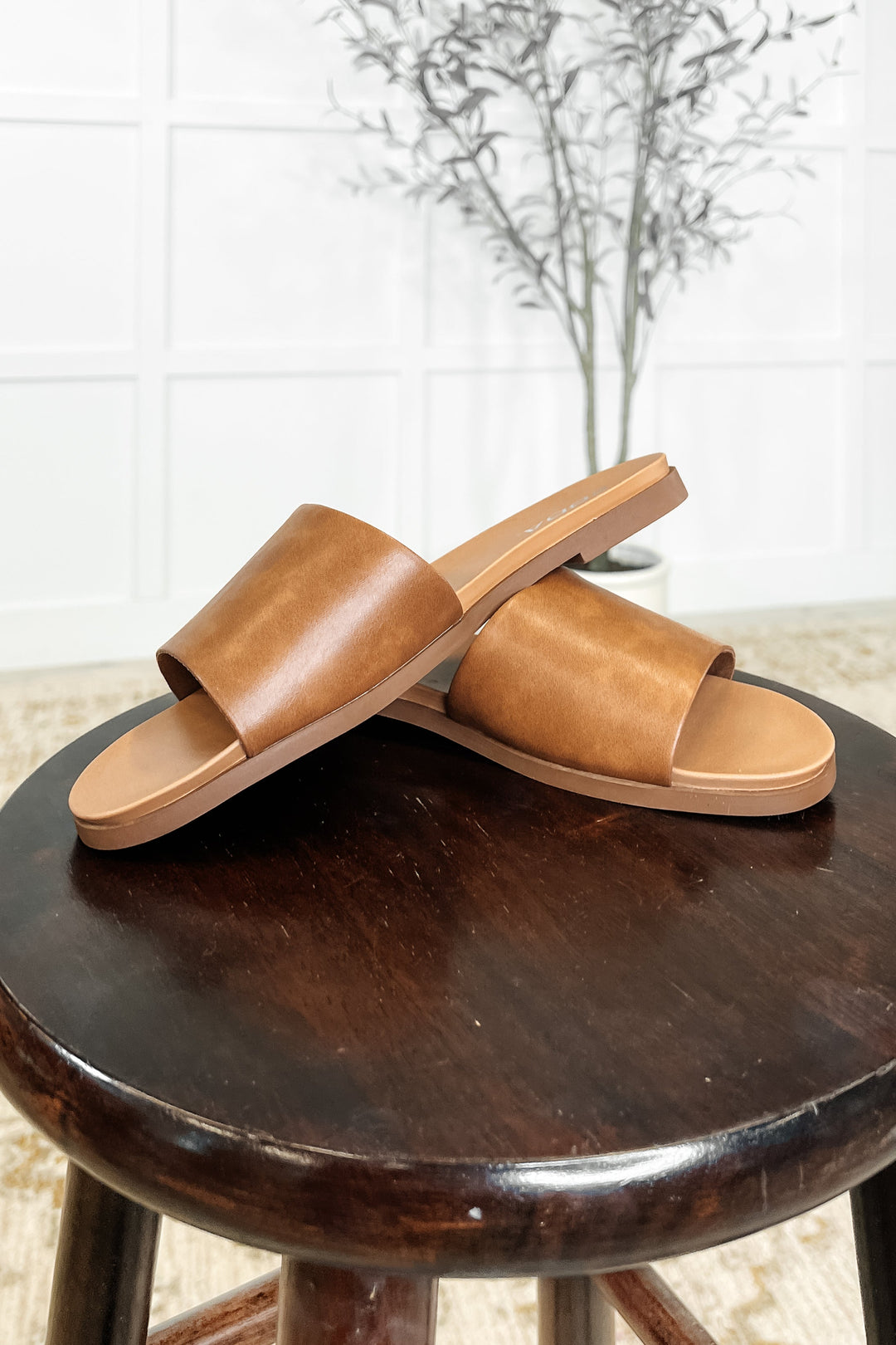 Pair of tan leather single-band slide sandals displayed on a dark wood stool with a neutral background and dried olive branch