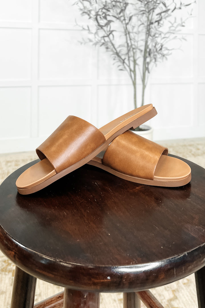 Pair of tan leather single-band slide sandals displayed on a dark wood stool with a neutral background and dried olive branch