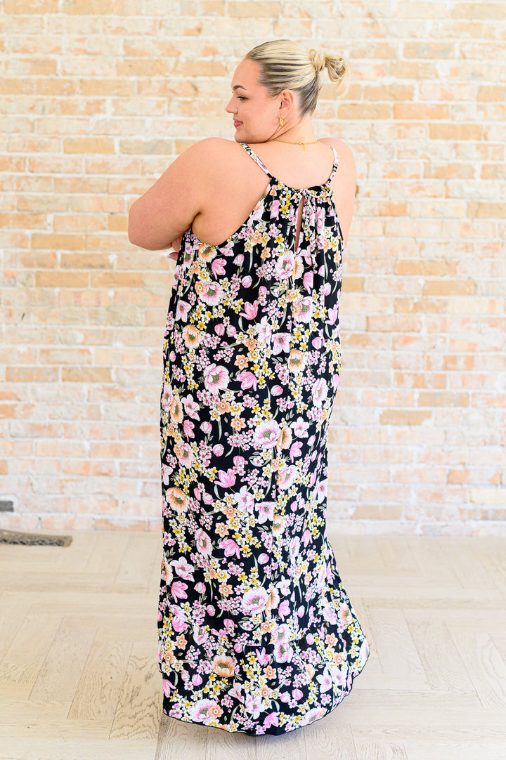 Back view of plus size model wearing a black floral midi dress showing halter neckline with tie detail and open back, styled with black flip flops.