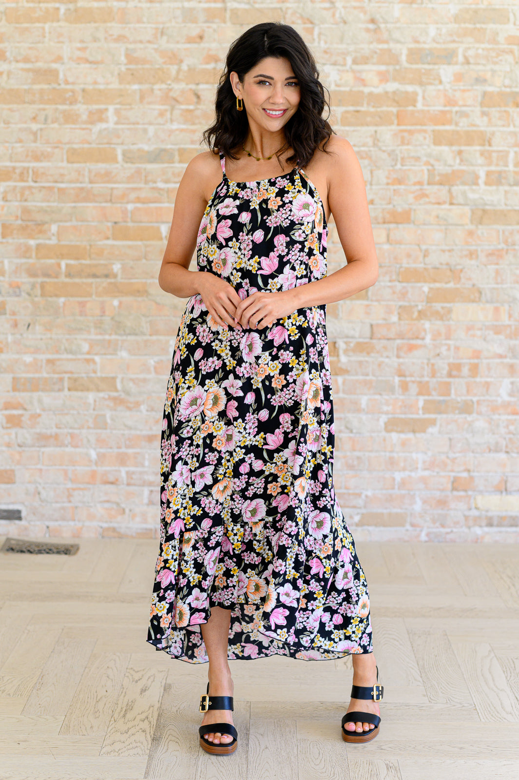 Woman wearing a black floral midi dress with pink, peach, and yellow blooms, spaghetti straps, and high-low ruffle hem, styled with black block heel sandals.