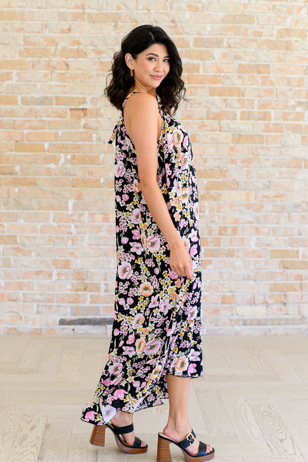Side view of woman wearing a black floral midi dress with spaghetti straps and high-low ruffle hem, styled with black block heel sandals.