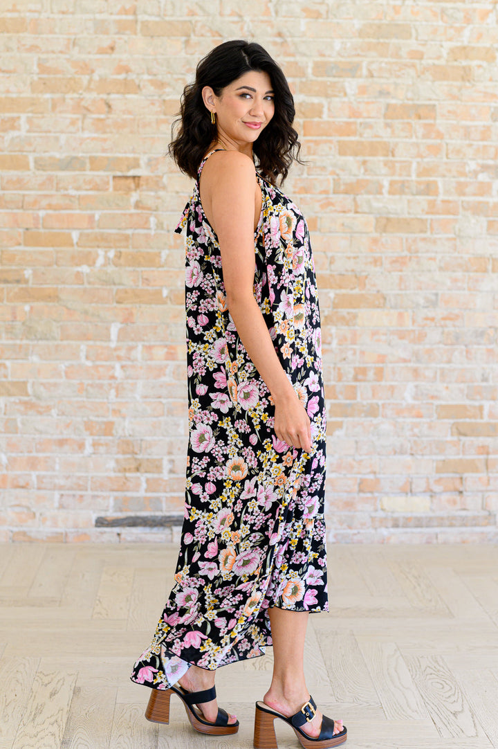 Side view of woman wearing a black floral midi dress with spaghetti straps and high-low ruffle hem, styled with black block heel sandals.
