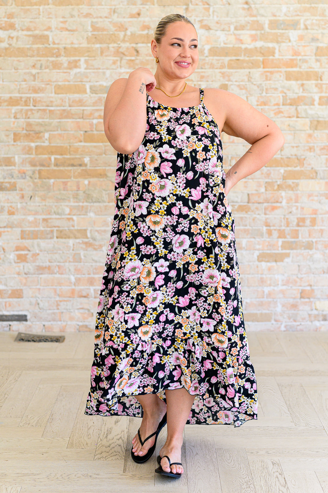 Plus size model wearing a black floral midi dress with pink, peach, and yellow blooms, spaghetti straps, and high-low ruffle hem, styled with black flip flops.