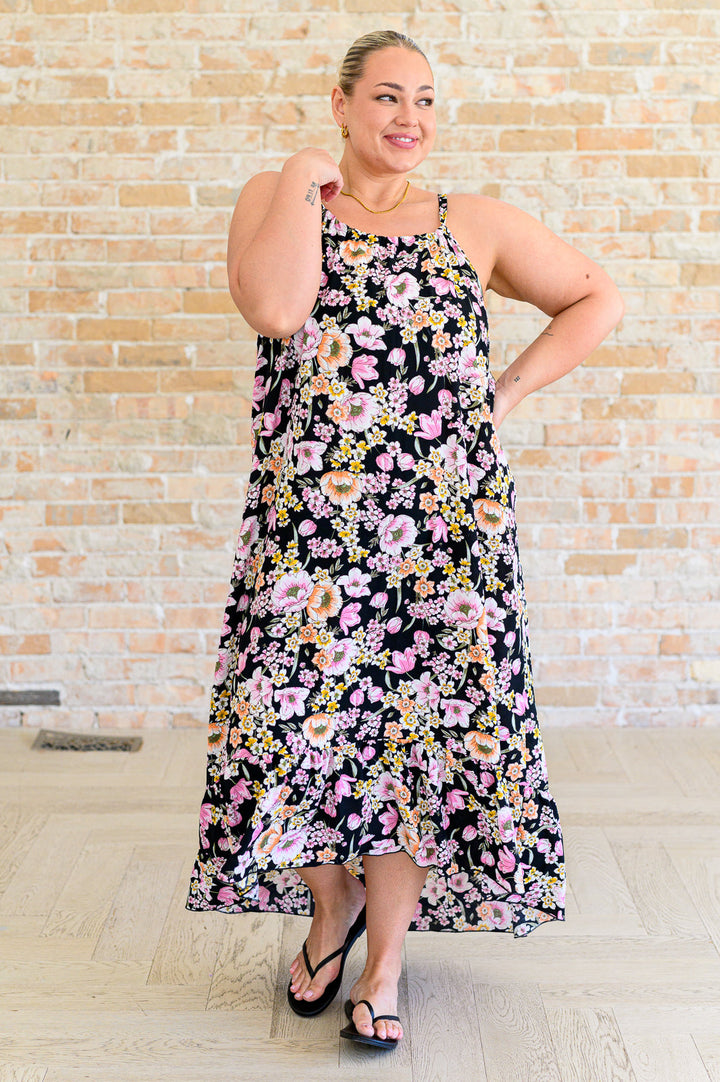 Plus size model wearing a black floral midi dress with pink, peach, and yellow blooms, spaghetti straps, and high-low ruffle hem, styled with black flip flops.