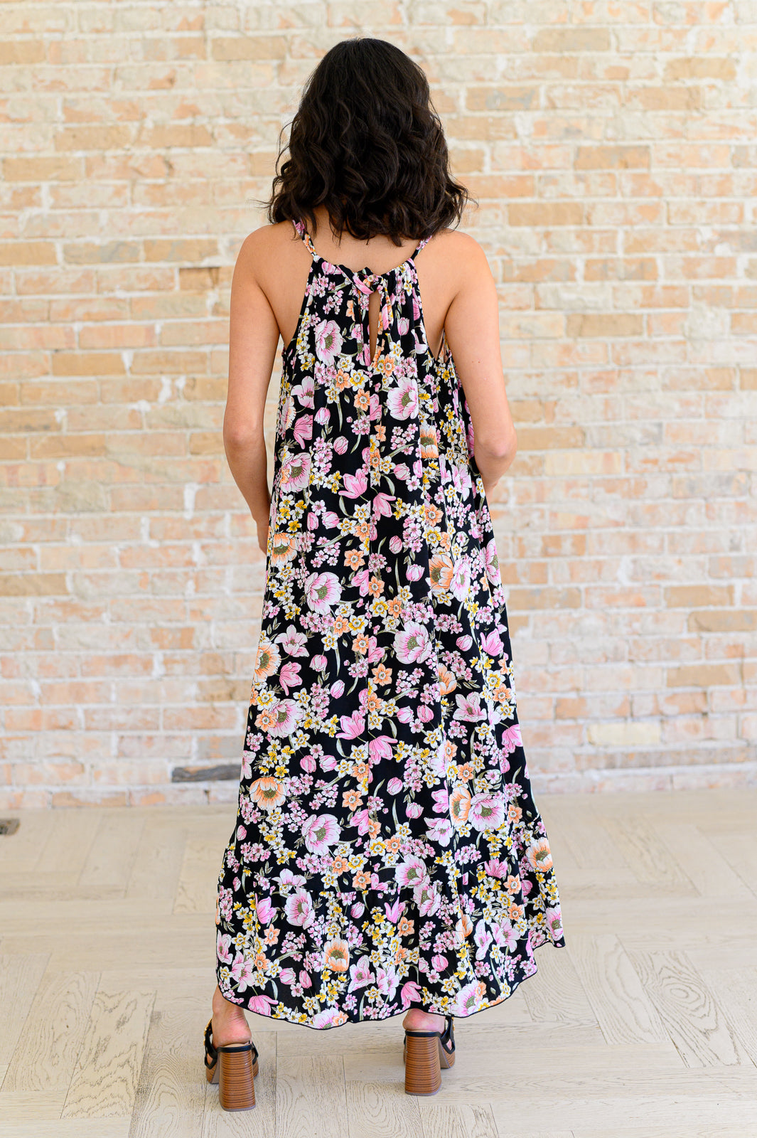 Back view of black floral midi dress showing halter neckline with tie detail, open back, and high-low ruffle hem, styled with black block heel sandals.