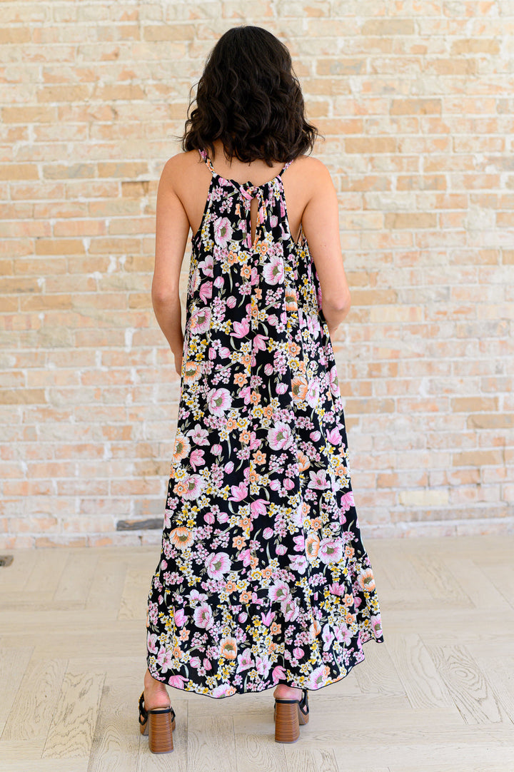 Back view of black floral midi dress showing halter neckline with tie detail, open back, and high-low ruffle hem, styled with black block heel sandals.
