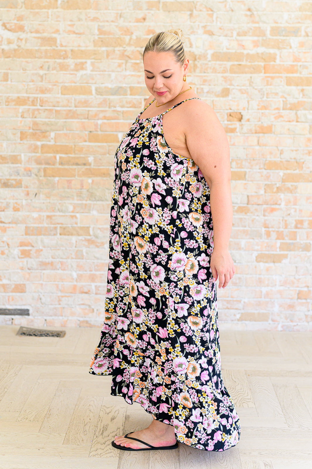 Side view of plus size model wearing a black floral midi dress with spaghetti straps and high-low ruffle hem, styled with black flip flops.
