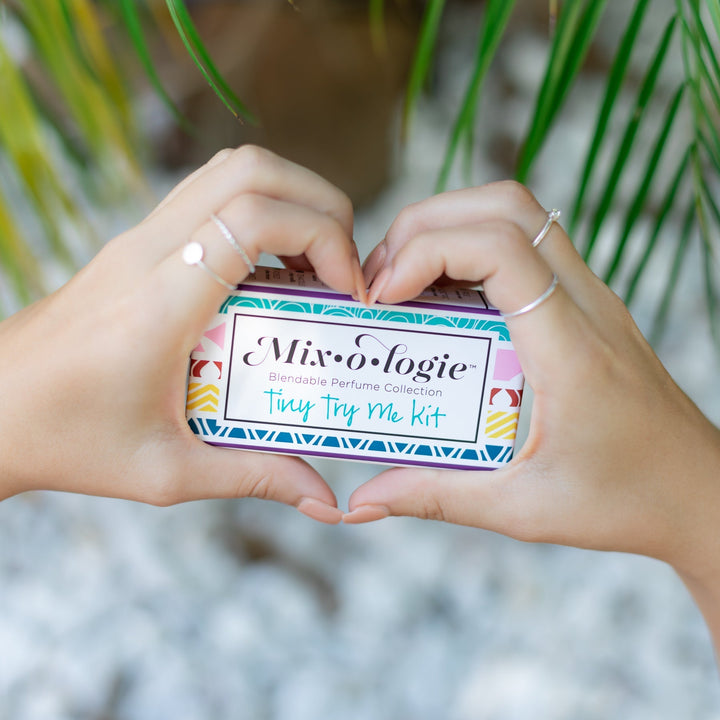 Mixologie Tiny Try Me Kit closed slide box held in heart shape by two hands with silver rings, with palm leaves and white pebbles in background